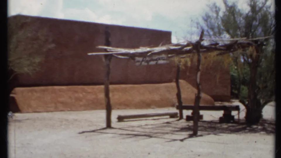 1971 California 8mm frame showing an adobe-style exterior, rustic shade arbor, bench, desert shrubs, and sunlit open ground.
