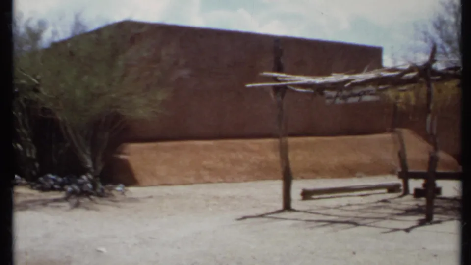 1971 California 8mm frame with an adobe-style wall, wooden shade structure, bench, desert planting, and bright sky.