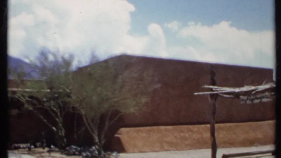 1971 California 8mm frame showing an adobe-style building, desert trees, a wooden arbor, distant mountains, and a partly cloudy sky.