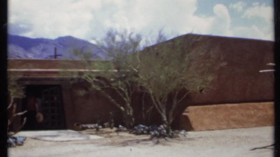 1971 California 8mm frame showing an adobe-style entrance, desert landscaping, arbor posts, distant mountains, and a possible person near the doorway.