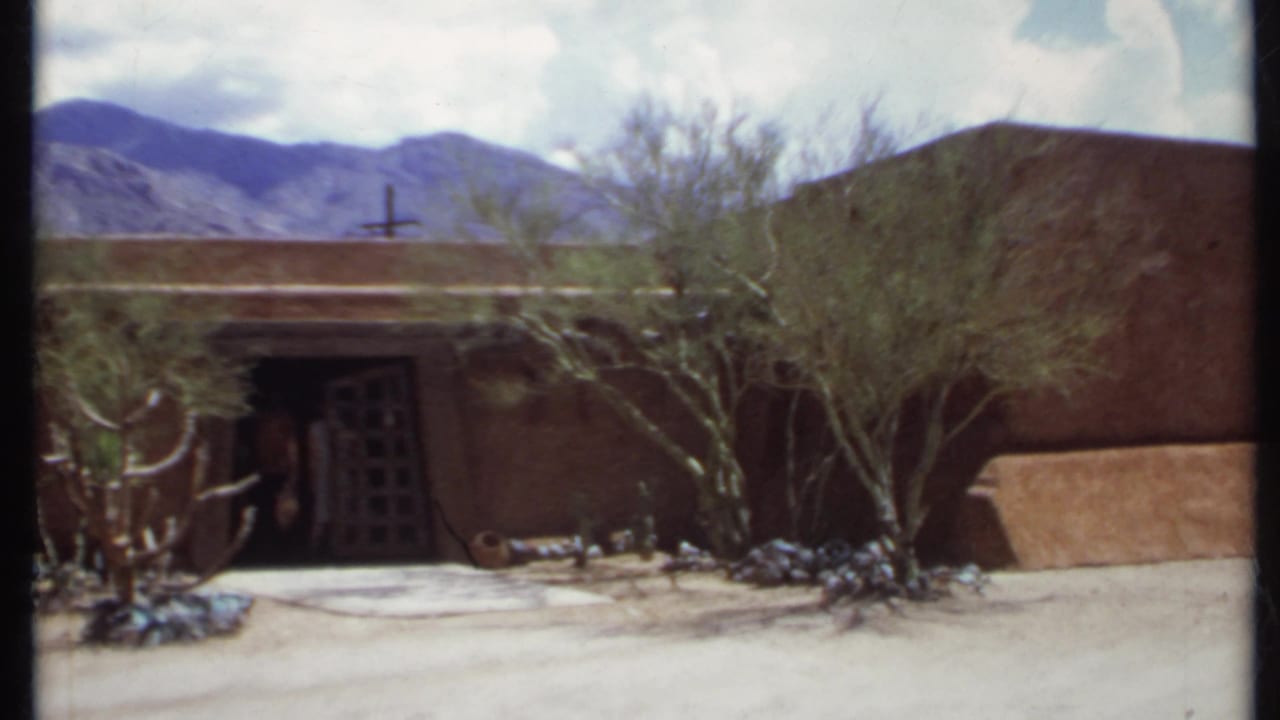 Adobe-like building with dark doorway, desert plants, mountains, and two small figures near the entrance.