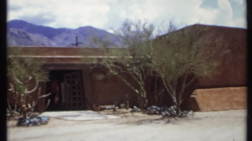 1971 California 8mm frame showing an adobe-like desert building, grid-pattern door, shrubs, cacti, mountains, and people near the entrance.