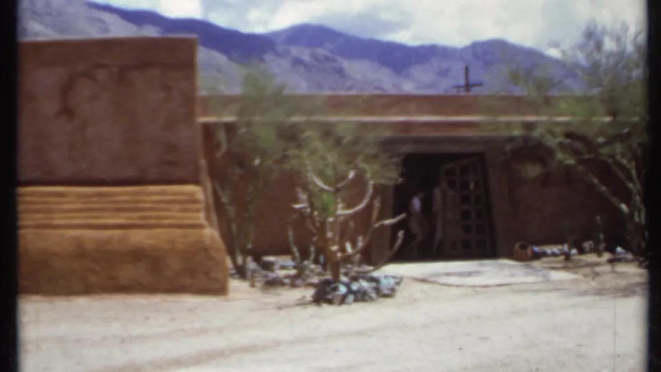 1971 California frame with an adobe-like exterior, stepped wall base, dark doorway, desert vegetation, mountains, and a small cross on the roofline.