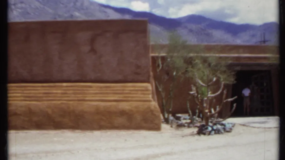 1971 California 8mm frame showing a closer adobe-like wall, stepped base, dark grid doorway, desert plants, mountains, and a person entering.