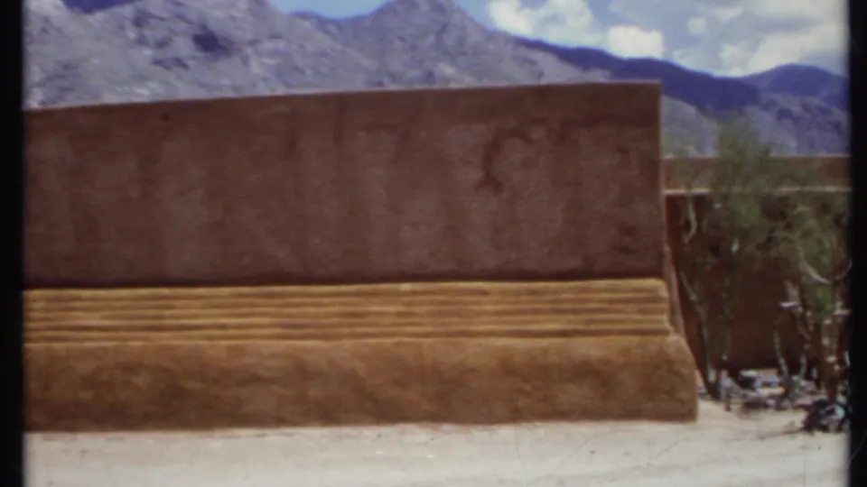 1971 California frame dominated by an adobe-like wall and stepped base, with desert plants, mountains, sky, and part of a doorway at right.