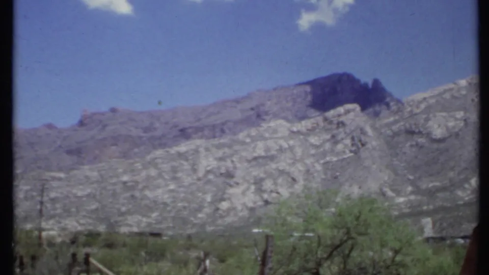 1971 California home-movie view of a rocky mountain ridge, sparse desert vegetation, and fence posts beneath blue sky.