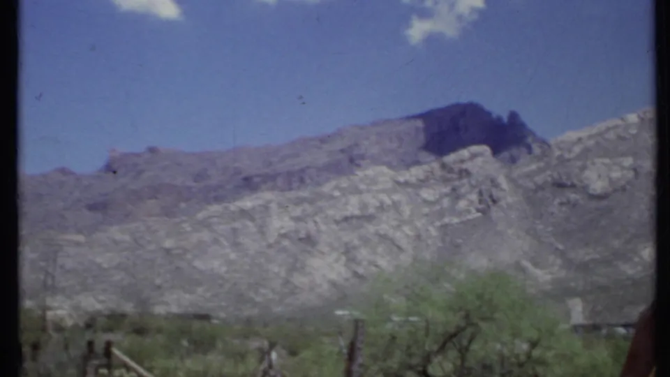 1971 California 8mm frame of a rugged mountain slope above sparse shrubs and fence posts in an arid landscape.