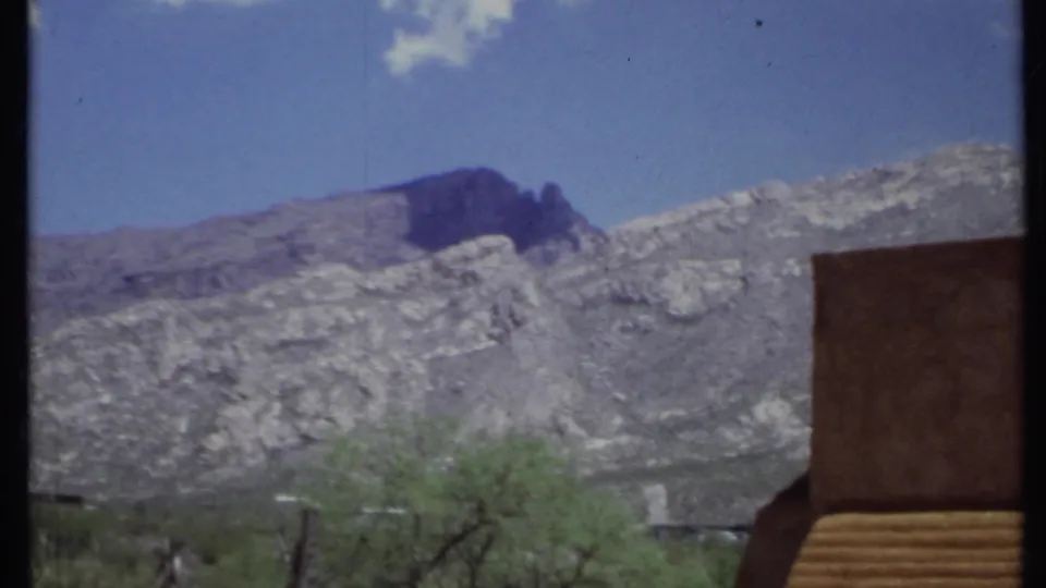 1971 California home-movie frame with rocky mountains, desert shrubs, and part of an adobe-style building at right.