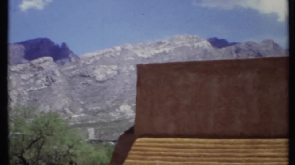 1971 California 8mm frame with a dry gray mountain ridge behind an adobe-style wall, steps, trees, and low desert vegetation.
