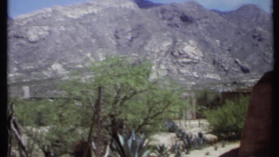 1971 California 8mm frame showing rocky mountains, desert trees, agave-like plants, fencing, and a small partially obscured structure.