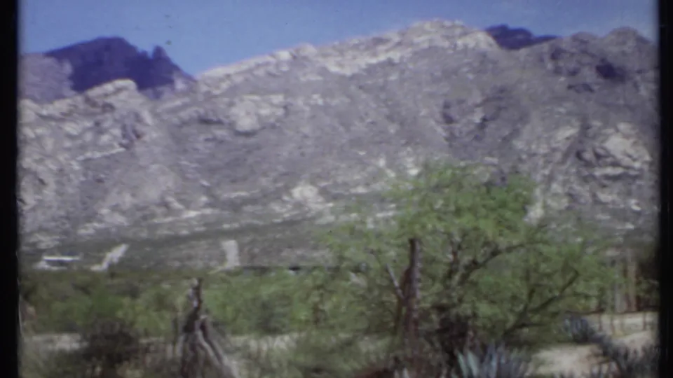 1971 California 8mm frame with a gray rocky mountain ridge, blue sky, desert trees, agave-like plants, and dry scrubland.