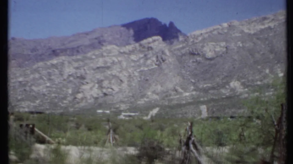 1971 California desert valley with gray mountain slopes, scattered shrubs, rough fencing, and small distant structures.