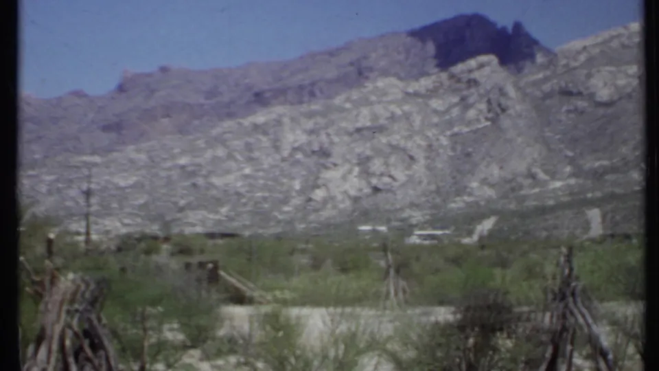 1971 California 8mm frame showing scrubby desert vegetation, rough posts, and a rocky mountain range under clear sky.