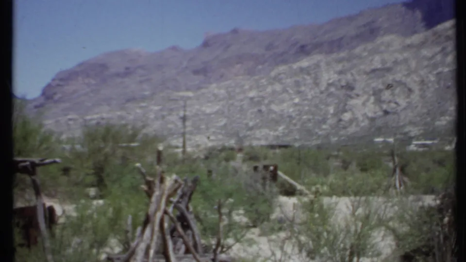 1971 California desert view with weathered wood posts, green scrub, distant small structures, and rocky mountains beyond.