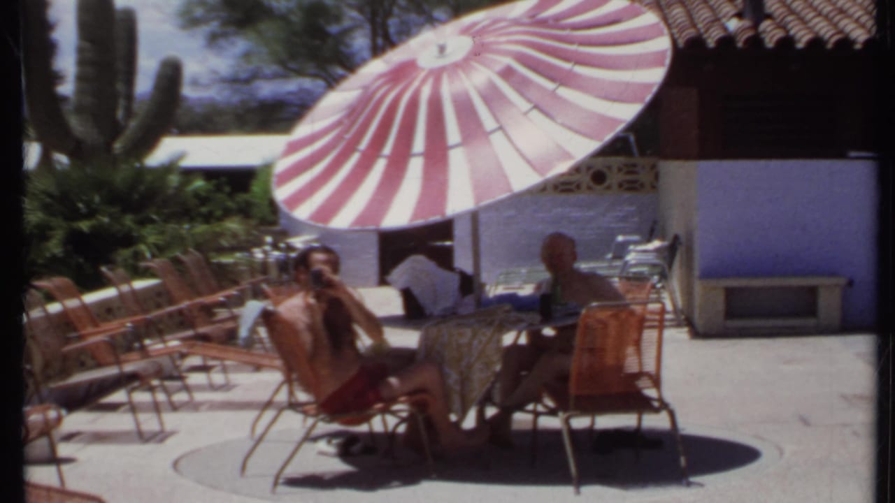 1971 California home-movie frame showing a man in red swim trunks photographing from a shaded patio table.
