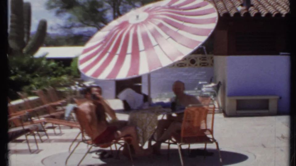 1971 California 8mm frame of two men seated in shade beside wicker patio furniture and desert planting.