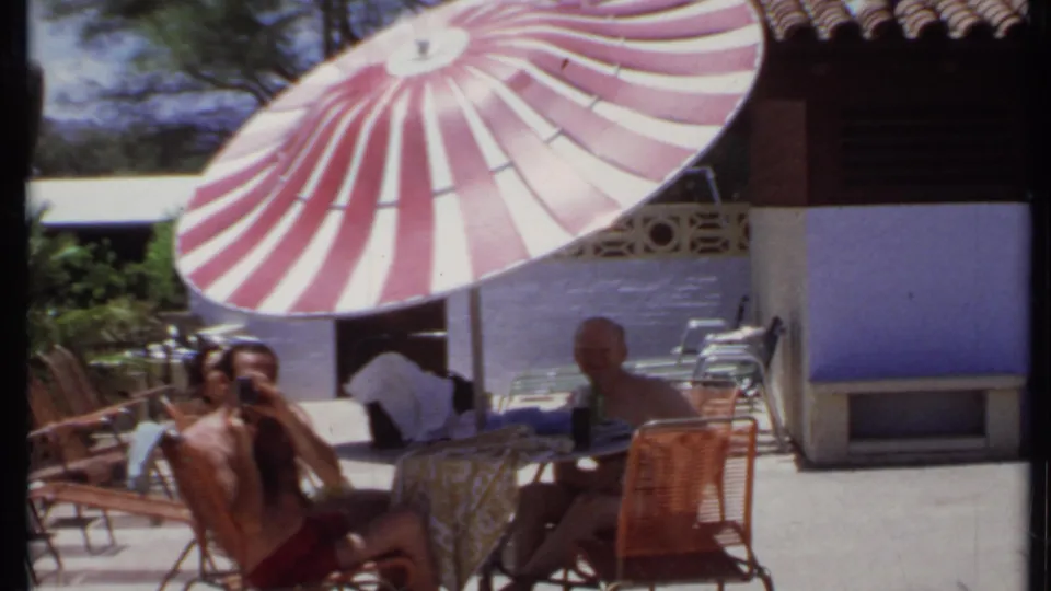 1971 California home-movie frame of two men seated under a striped umbrella beside patio furniture and a stucco building.