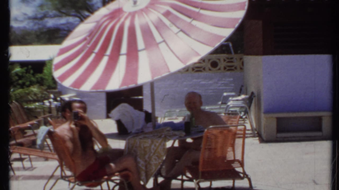 Two men sit at a patio table beneath a red and white striped umbrella.