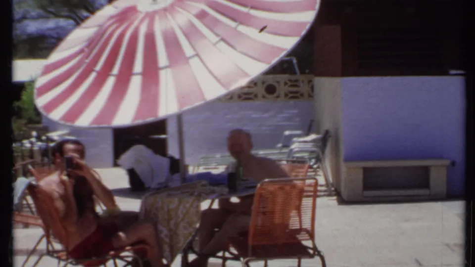 1971 California patio scene with two adult men seated at a table under a red and white striped umbrella near a purple wall.