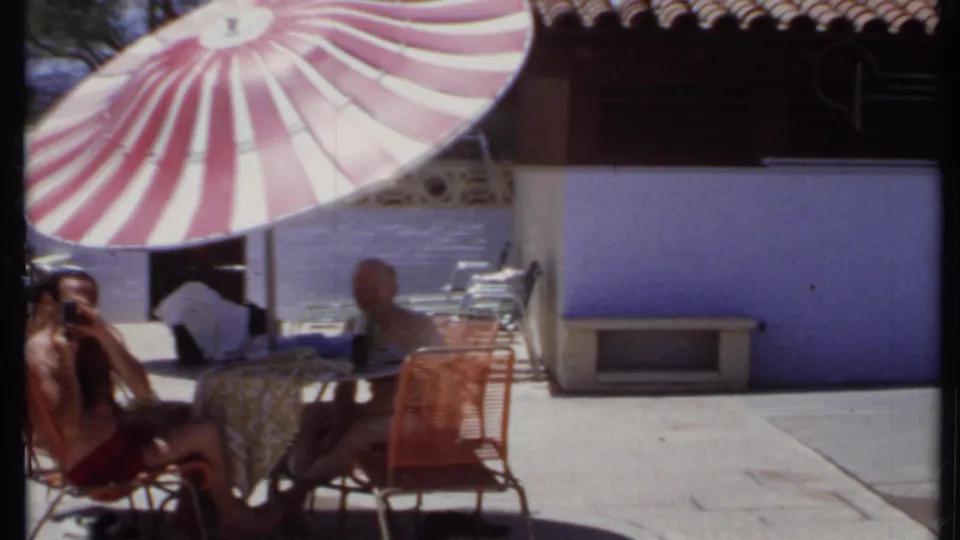1971 California 8mm frame with two men partly framed at a patio table under a large striped umbrella.