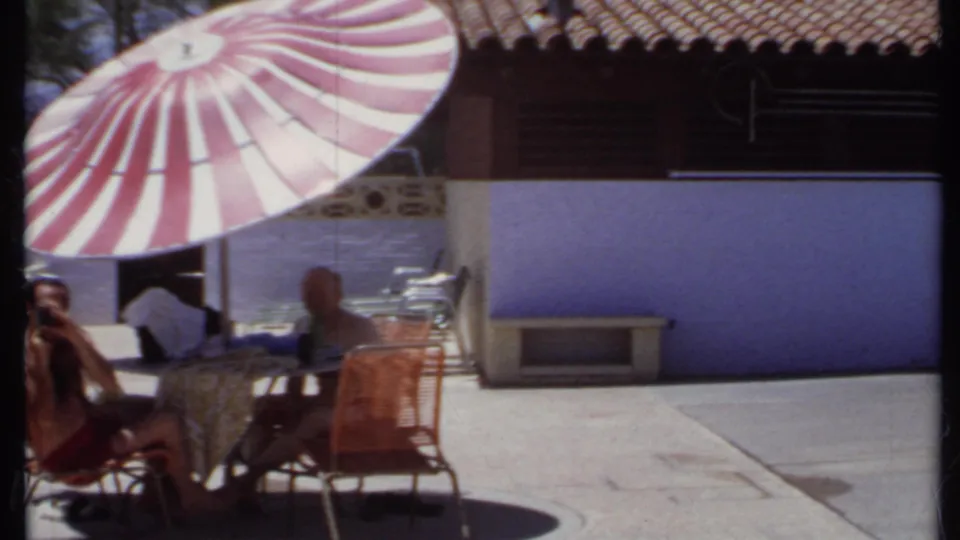 1971 California home-movie frame of a patio with two seated men, wicker furniture, and a red and white umbrella against a purple wall.