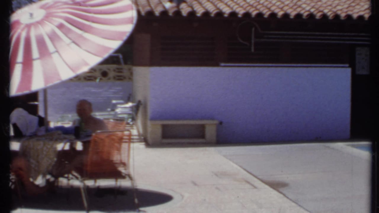 Shaded patio table beside a pool with a red-and-white umbrella and tiled-roof building.
