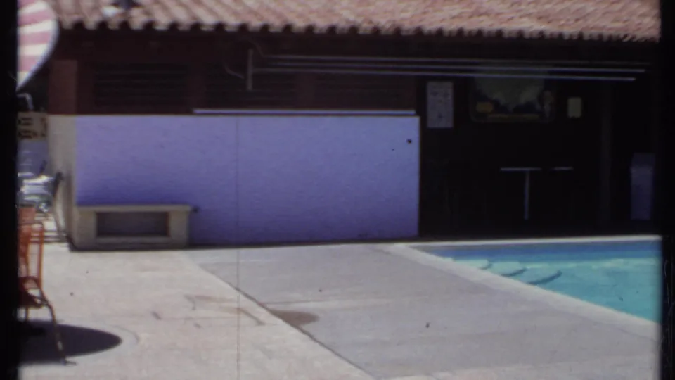 California poolside view from 1971 with a purple wall, tiled-roof structure, umbrella edge, and turquoise pool water.
