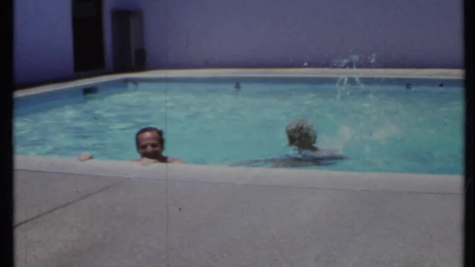 1971 California home-movie frame of two adult swimmers in a pool, one wearing a patterned swim cap.