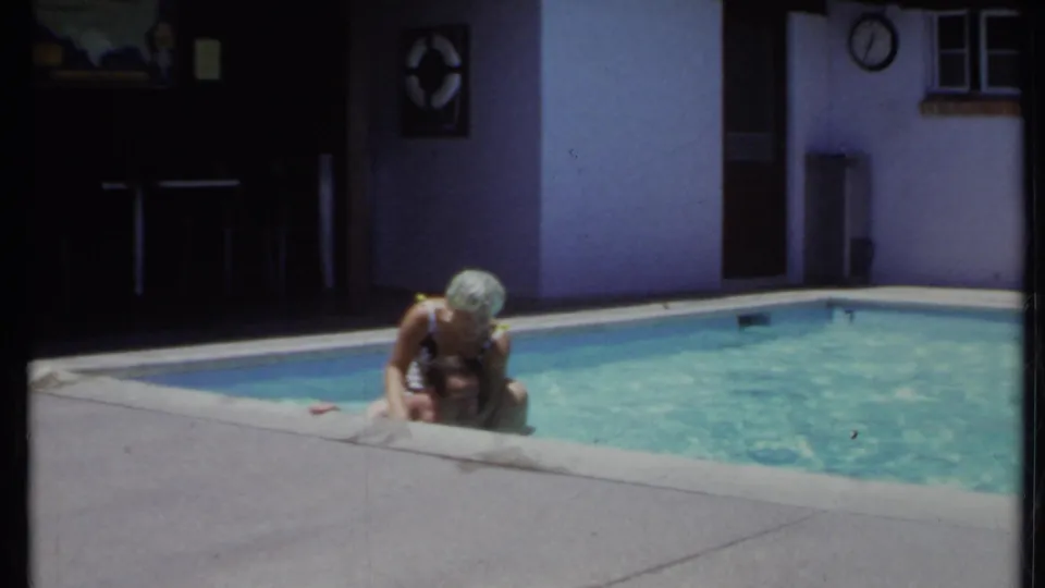 1971 California home-movie frame with a woman in a patterned swim cap leaning over a shirtless man in a backyard pool.