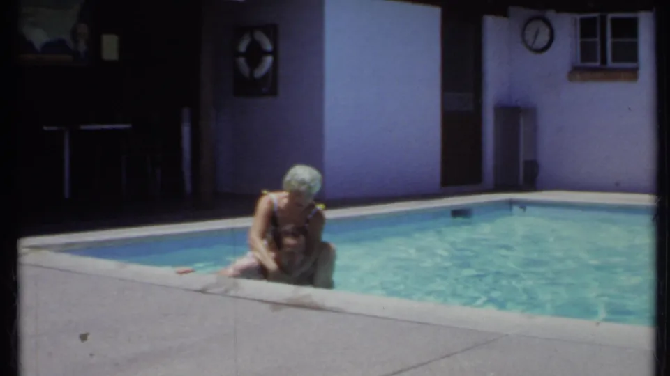 1971 California 8mm frame showing a woman in a patterned swim cap and dark swimsuit leaning over a man near the pool coping.
