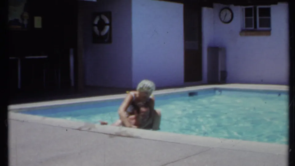 1971 California home-movie frame of two adults at a backyard pool edge, with stucco walls, a wall clock, and bright water visible.