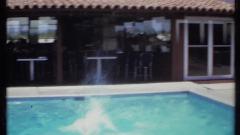 1971 California 8mm frame showing a turquoise backyard pool beside a stucco house with tiled roof and covered patio seating.