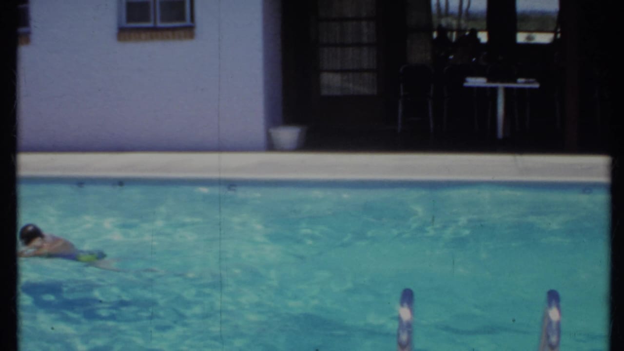 1971 California home-movie frame of swimmers moving through a backyard pool in front of a stucco house and shaded patio.