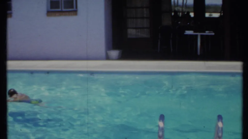 1971 California home-movie frame of swimmers moving through a backyard pool in front of a stucco house and shaded patio.