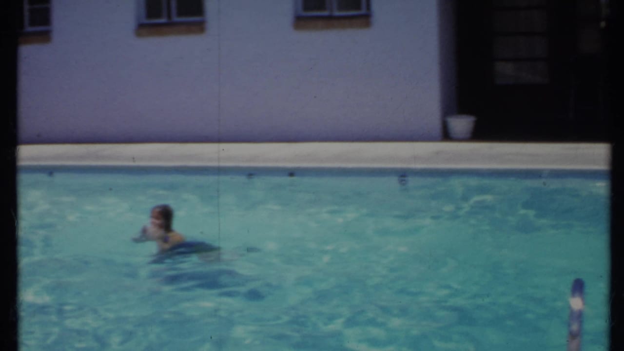 1971 California 8mm frame showing a swimmer in a blue flotation ring in a backyard pool beside a stucco house wall.