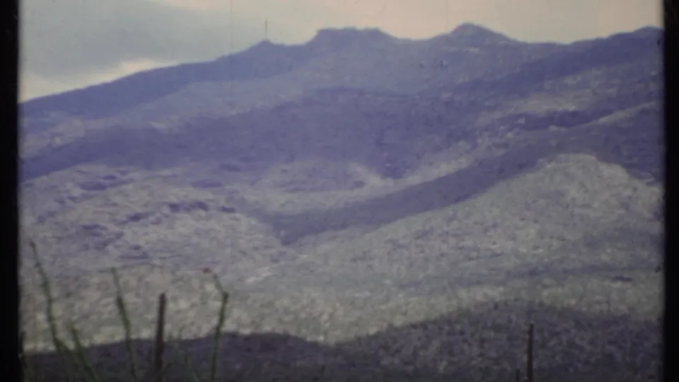 1971 California 8mm frame showing rugged mountain ridges, muted color, and thin desert plants at the lower edge.