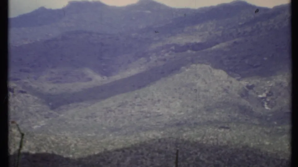 1971 California home-movie frame with blue-purple mountain slopes, pale sky, and visible 8mm gate edge down the center.