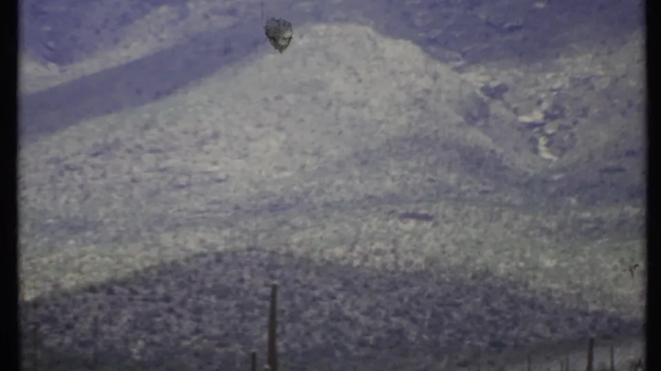 1971 California home-movie frame of a rugged mountain slope with sparse desert vegetation, grain, and small film blemishes.