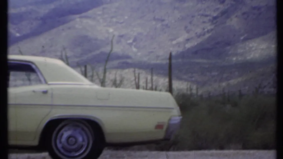 1971 California home-movie frame showing a parked light-colored sedan near cacti below rugged desert mountains.