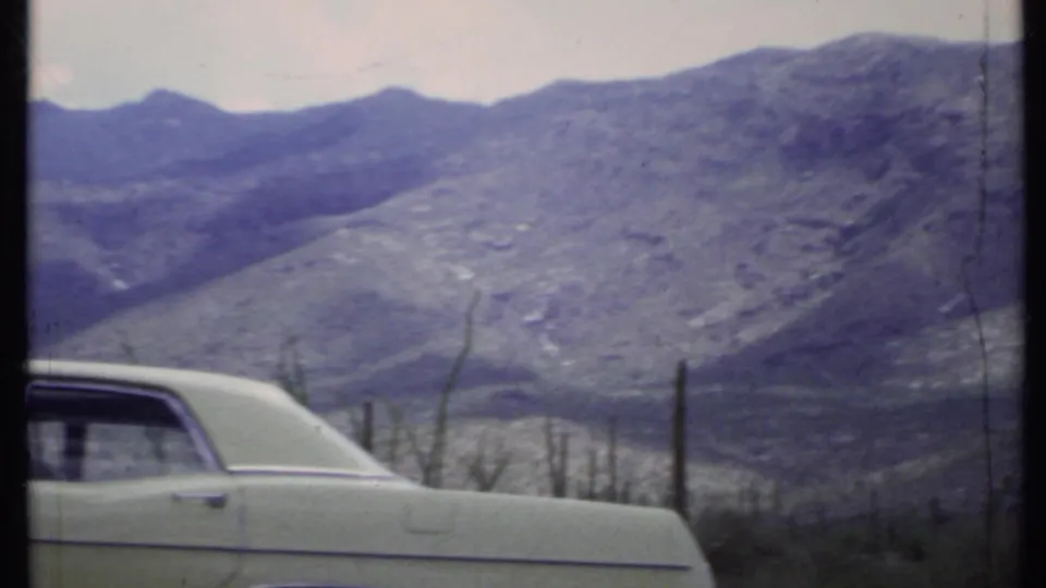 1971 California desert frame with a parked light-colored sedan in the foreground and mountain ridges beyond.