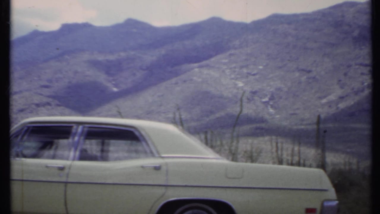 1971 California home-movie frame showing a pale sedan in the foreground with rugged desert mountains and cactus-like vegetation behind it.
