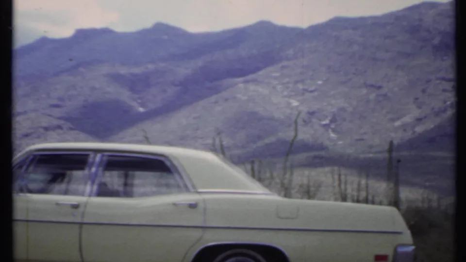1971 California home-movie frame showing a pale sedan in the foreground with rugged desert mountains and cactus-like vegetation behind it.