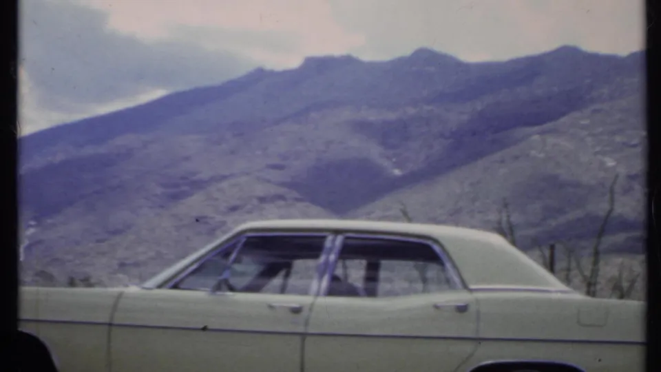 The sedan is framed from the side against arid California mountain slopes, with faint occupants visible through the windows.