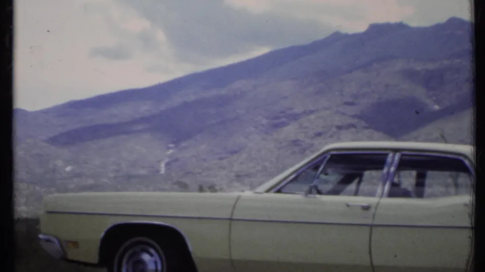 A 1971 home-movie frame shows the front portion of a pale sedan beside desert scrub, with mountains filling the background.
