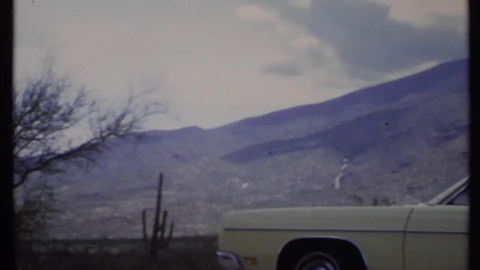 1971 8mm California desert road-trip frame with a pale yellow car, tall cactus, bare trees, and distant mountains under clouds.