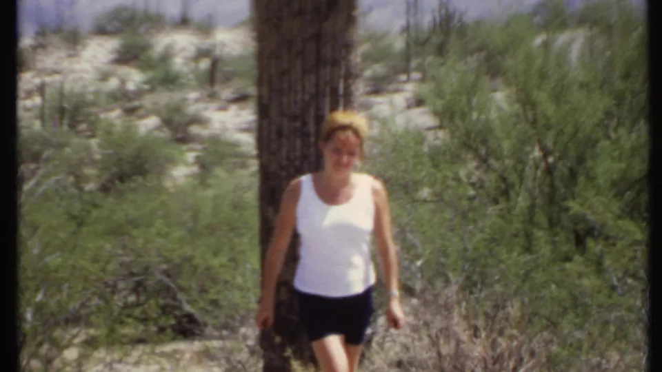 Adult woman in a white sleeveless top and dark shorts walks through a 1971 California desert setting near cactus and green brush.