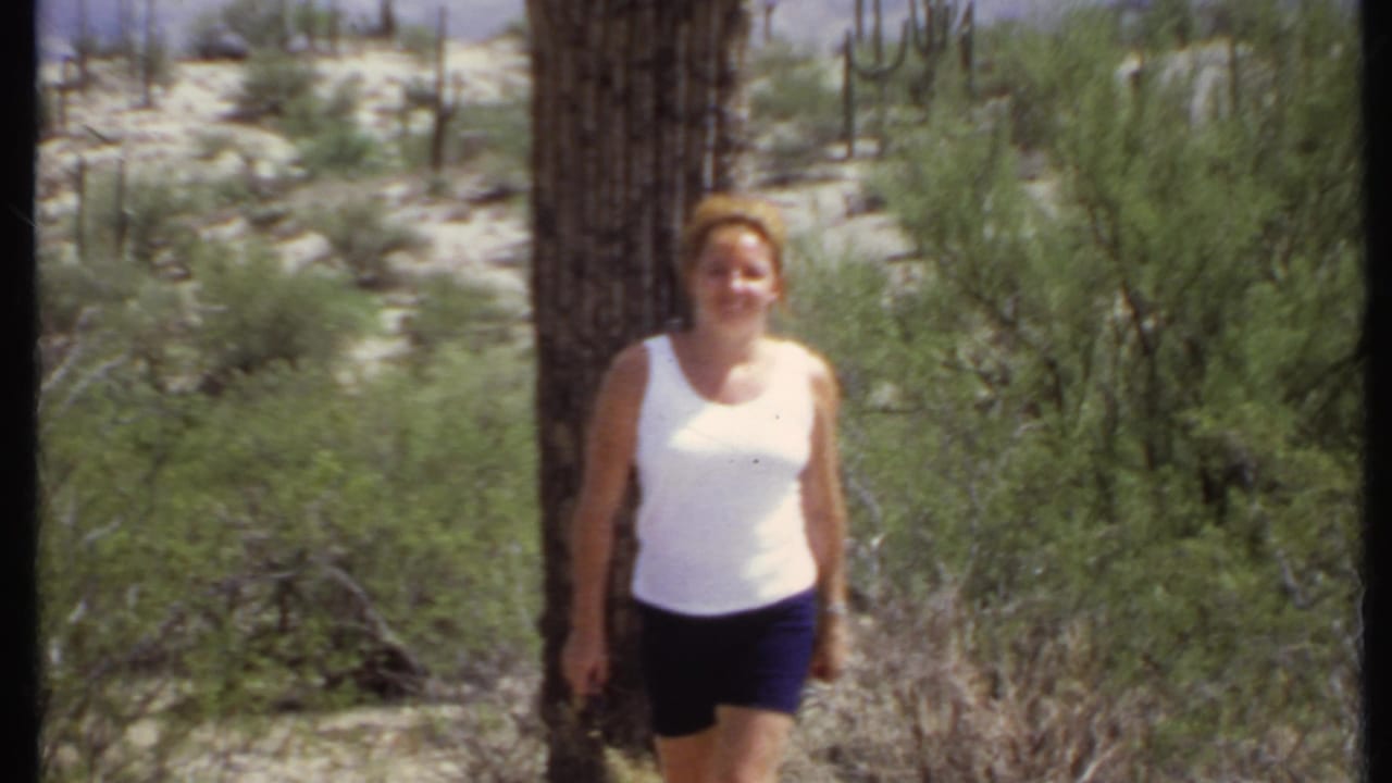 1971 home-movie frame of a woman in desert vegetation near a tall cactus, with pale ground and shrubs behind her.