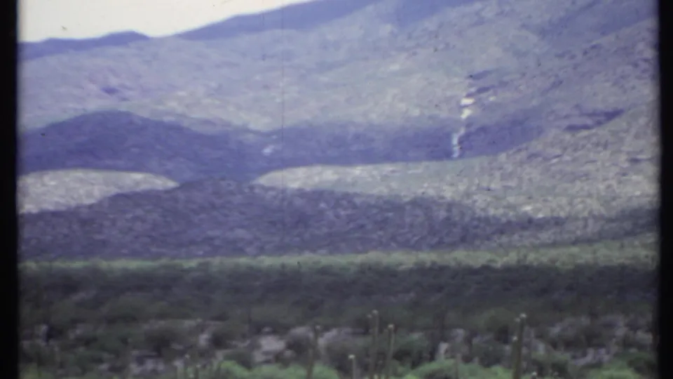 1971 California home-movie frame showing distant arid mountains, a green valley band, and cactus tops along the lower edge.