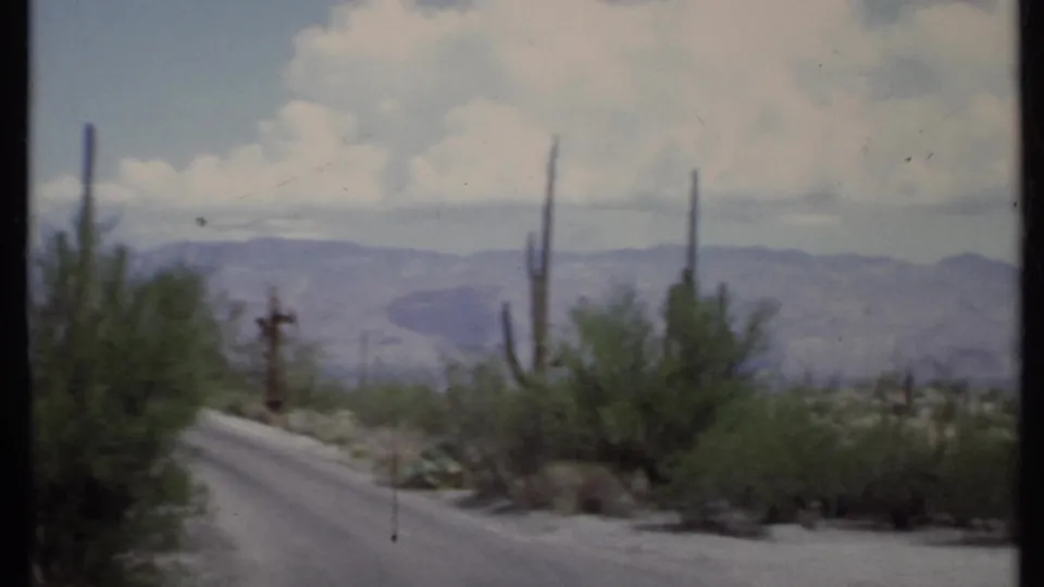 1971 8mm California desert road with tall cacti, roadside scrub, mountains, and visible film-frame edges.