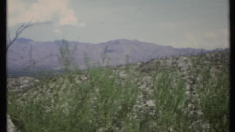 1971 California 8mm frame with desert shrubs in the foreground, a rocky rise, distant mountains, and soft clouds overhead.